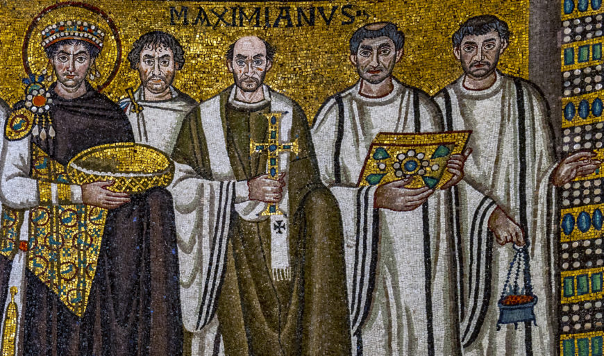 Detail of a mosaic artwork depicting a group of five robed figures, with one holding a golden bowl and another holding a decorated book, set against a gold background with the inscription "MAXIMIANVS." The image highlights intricate details of clothing, accessories, and religious symbols, emphasizing historical and religious significance.