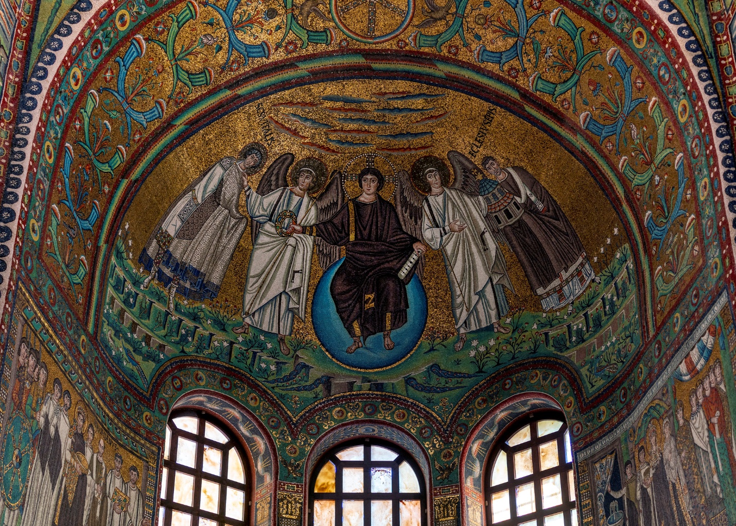 Photograph of a detailed Byzantine-style mosaic inside a domed ceiling, depicting a central figure seated on a blue orb surrounded by four angels. The mosaic features vibrant gold, green, and blue colors with intricate patterns and three arched windows below.