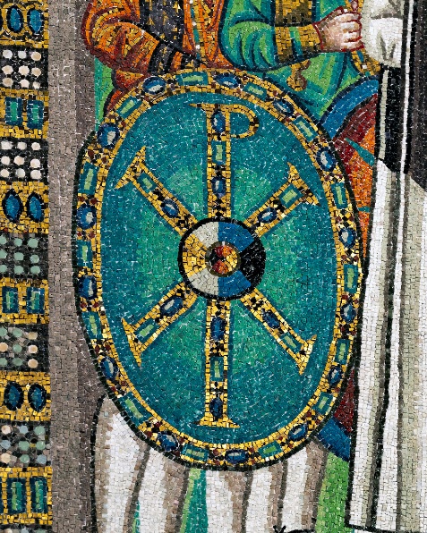 Detail of a mosaic artwork depicting a detailed oval shield with a Chi-Rho symbol in gold and blue on a turquoise background. The shield is held by a figure dressed in colorful garments, with intricate patterns and textures visible in the surrounding mosaic tiles.