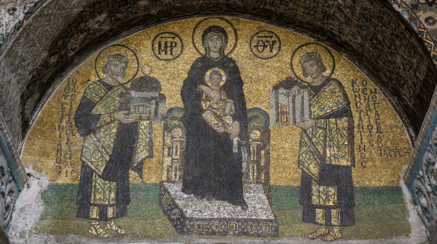A mosaic artwork depicting a central seated figure holding a child, flanked by two standing figures presenting architectural models. The mosaic features gold backgrounds, intricate patterns, and Greek inscriptions above and beside each figure.