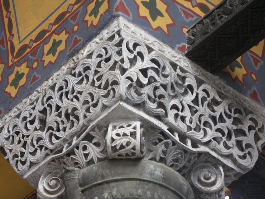 Photograph of an intricately carved stone architectural detail featuring swirling, leaf-like patterns on a corner capital. Surrounding elements include colorful painted walls with geometric star motifs and a dark wooden beam with carved designs.