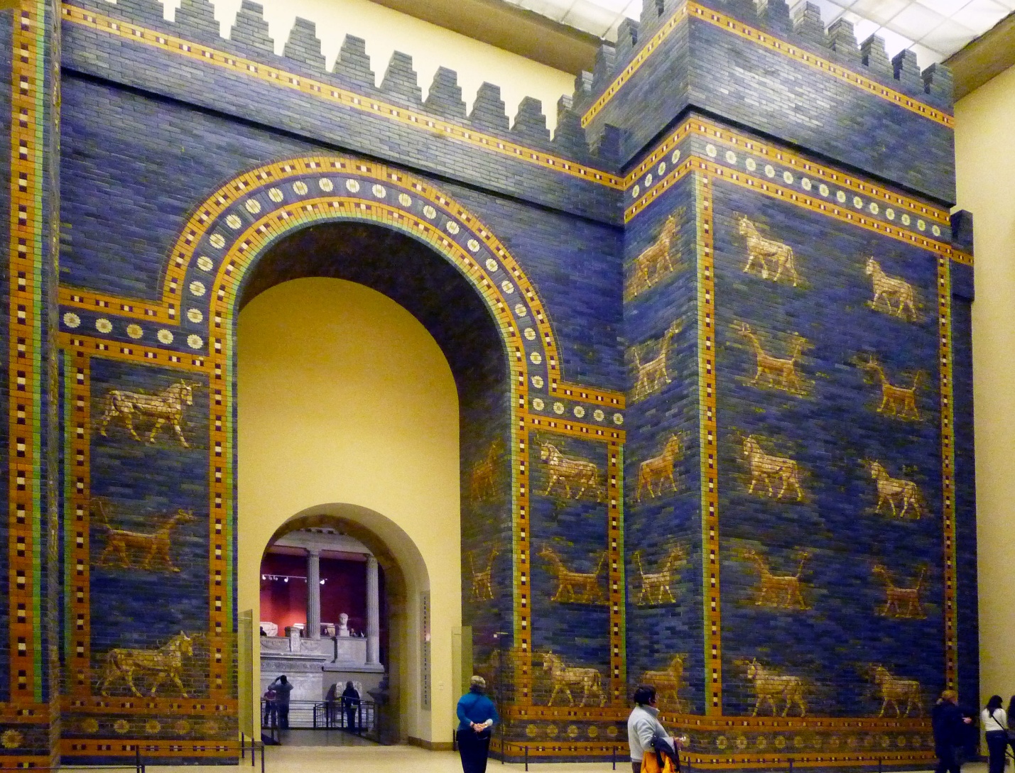 Photograph of the Ishtar Gate, an ancient Mesopotamian structure featuring deep blue glazed bricks decorated with golden animal reliefs, including lions, bulls, and dragons. The gate's large arched entrance and crenellated top are visible, with visitors providing scale inside a museum setting.