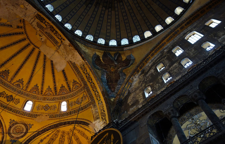 Photograph of an ornate interior ceiling featuring a large central dome with circular windows and intricate yellow and black geometric patterns. The image highlights architectural details including arches, columns, and a decorative emblem at the dome's base.