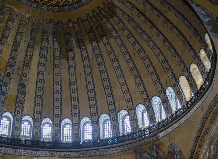 Photograph of an ornate dome ceiling featuring a series of tall, narrow arched windows with intricate blue and gold decorative patterns on ribs extending from the base to the apex. The design includes detailed geometric and floral motifs, with light streaming through windows.