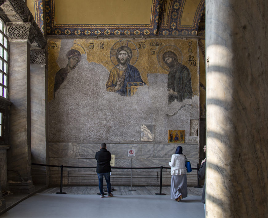 Photograph of a historic Byzantine mosaic inside a religious site, showing three partially damaged haloed figures with Greek inscriptions above them. Two visitors stand behind a barrier observing the mosaic, which features gold and blue colors with intricate border patterns on a high wall.