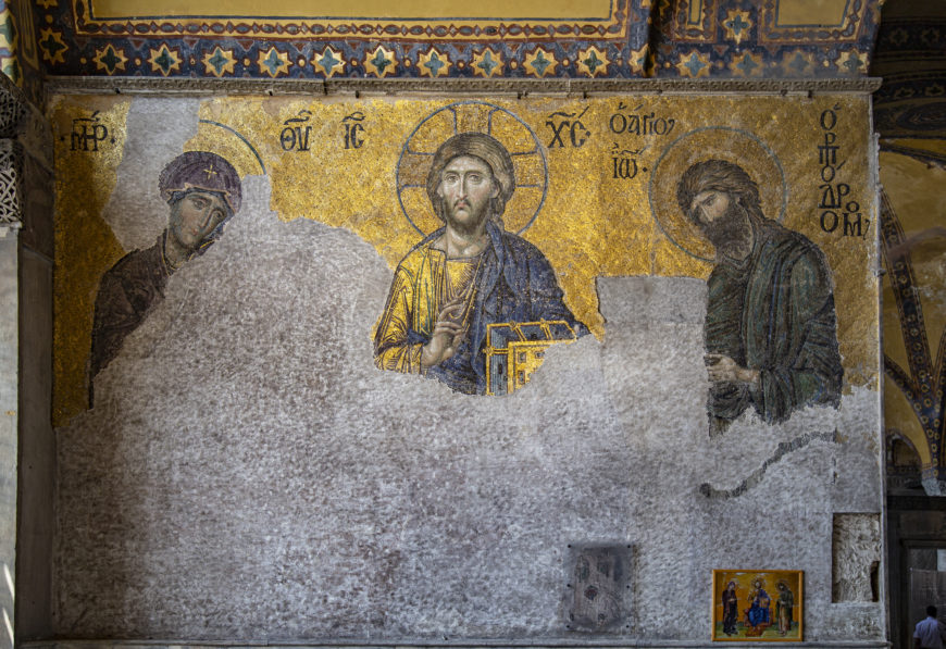 A photograph of a Byzantine mosaic depicting religious figures with halos, including a central figure holding a book and making a blessing gesture, set against a gold background. The mosaic shows significant wear and damage, with large portions missing or faded, and includes Greek inscriptions above each figure.
