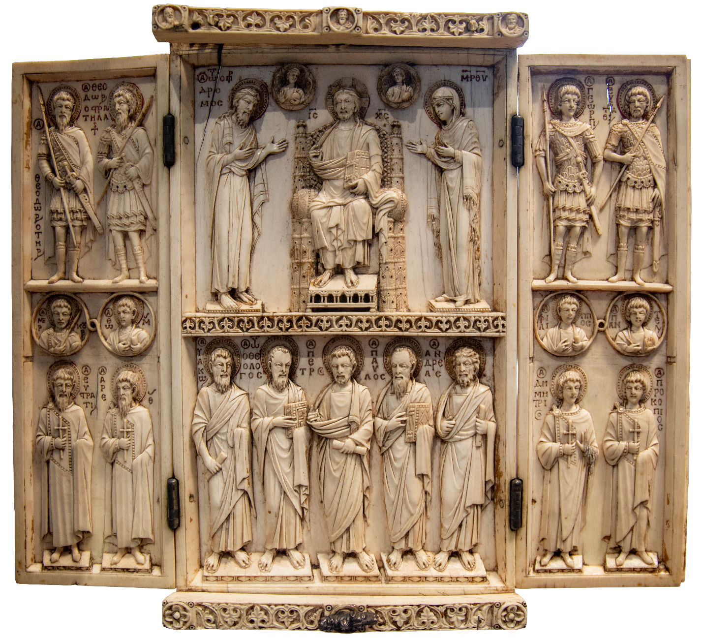 Triptych ivory carving depicting religious figures in a Byzantine style, with a central panel featuring a seated figure flanked by two standing figures and a lower row of six standing figures. Side panels contain pairs of standing saints and circular medallions with busts, all surrounded by intricate decorative borders and Greek inscriptions.