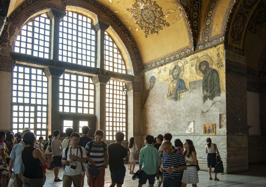 Photograph of a historic interior space with large arched windows and a mosaic depicting religious figures on a wall. The scene shows a group of diverse tourists observing and photographing the artwork under ornate ceiling decorations.