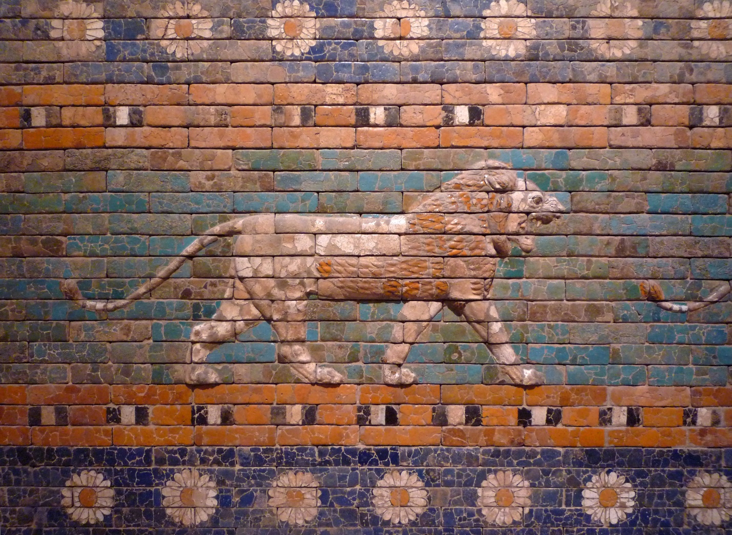 Photograph of Detail, Ishtar Gate and Processional Way brick relief depicting a lion walking to the right, set against a blue and green background with floral patterns above and below. The relief features detailed carving and color accents in orange, white, and black, highlighting the lion's mane and body structure.