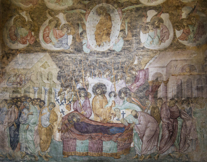 A fresco painting depicting a religious scene with a central figure surrounded by apostles and other figures, likely illustrating a significant biblical event. The artwork features muted earth tones, architectural elements, and a group of angels above, emphasizing the spiritual and ceremonial context.