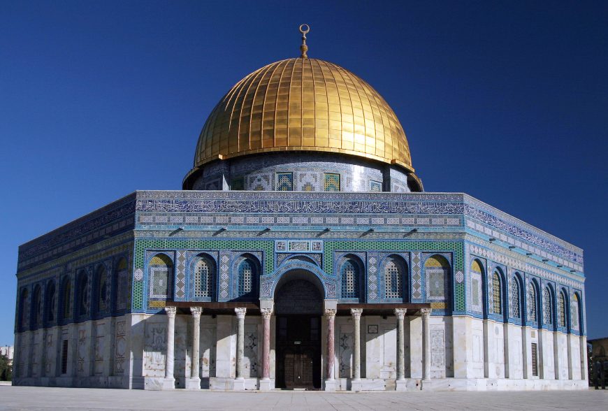 Photograph of Dome of the Rock, an Islamic shrine in Jerusalem, featuring a large golden dome atop a rectangular base adorned with intricate blue and green tilework and arched windows. The structure stands against a clear blue sky.