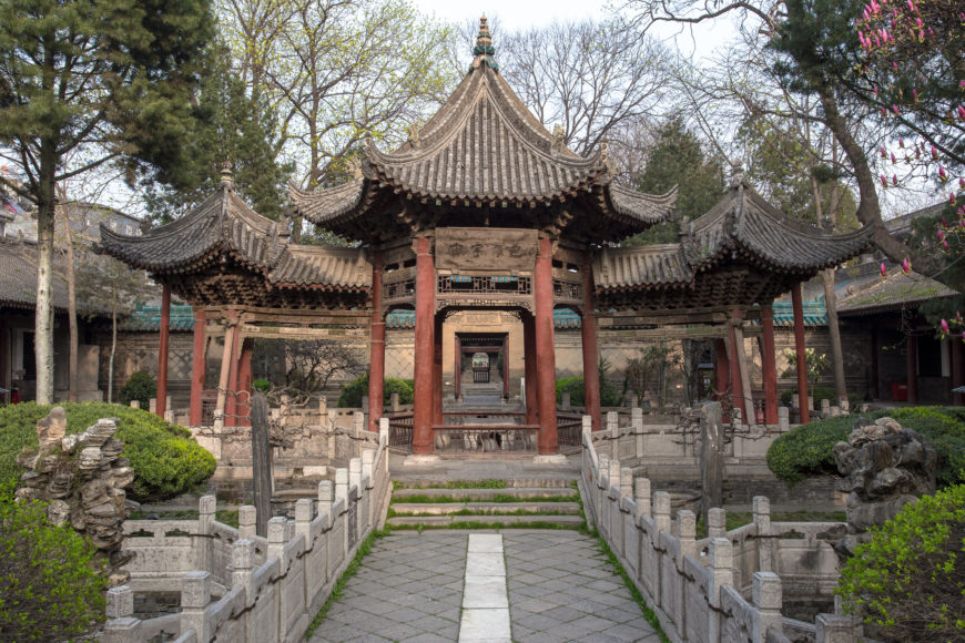 Photograph of a traditional Chinese pavilion with ornate, curved roofs supported by red wooden pillars, situated in a garden with stone pathways and greenery. The structure features intricate architectural details and is framed by trees, showcasing classical Chinese garden design and cultural heritage.
