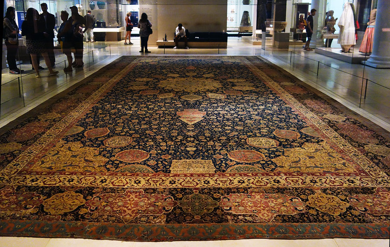 Photograph of an ornate Persian carpet displayed in a museum setting with visitors in the background. The carpet features intricate floral and geometric patterns in dark blue, red, and gold tones, highlighting traditional craftsmanship and cultural heritage.