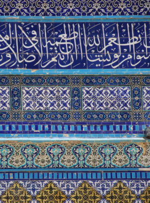 Photograph of intricate Islamic tilework featuring Arabic calligraphy in white on a deep blue background at the top, followed by geometric and floral patterns in blue, turquoise, and yellow hues arranged in horizontal bands. The design showcases traditional craftsmanship with detailed motifs and vibrant colors.