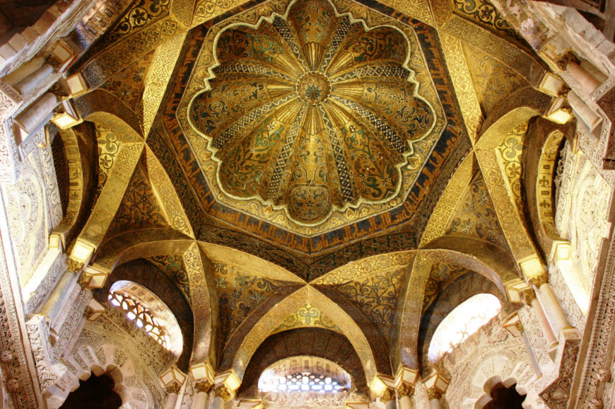 Photograph of an ornate, golden domed ceiling featuring intricate geometric and floral patterns with detailed arches supporting the structure. The design highlights Islamic architectural elements with symmetrical motifs and rich decorative artistry.
