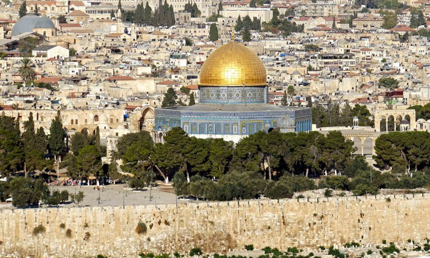 Photograph of the Dome of the Rock, an Islamic shrine with a golden dome and intricate blue tilework, situated in Jerusalem. The structure is surrounded by dense greenery and ancient stone walls, with a densely built cityscape in the background.