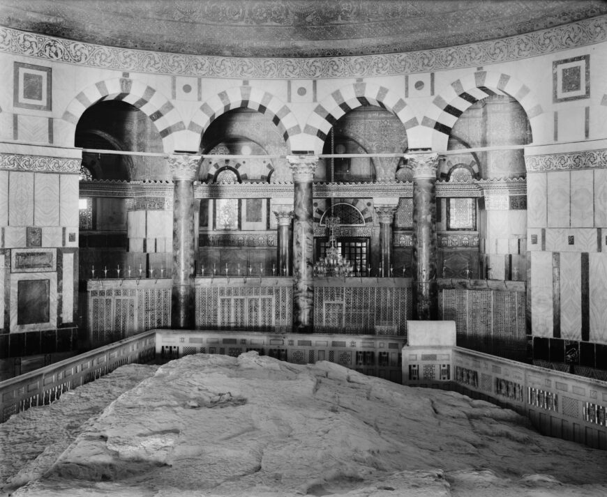 Photograph of Interior of the Dome of the Rock featuring a large stone slab surrounded by a decorative railing. The background showcases a series of arches with alternating light and dark stone patterns, intricate tile work, and detailed columns, highlighting historical architectural design.