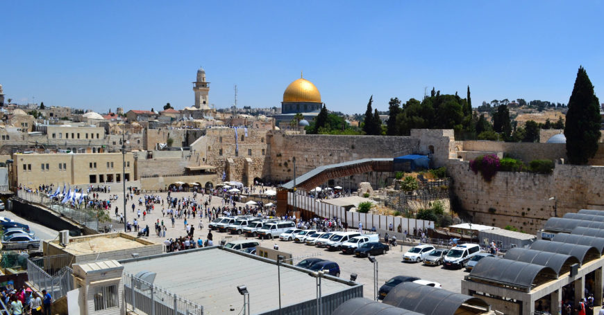 Photograph of a crowded plaza at the Western Wall in Jerusalem, featuring numerous visitors and parked cars. Prominent elements include the golden Dome of the Rock in the background and ancient stone walls surrounding the area.