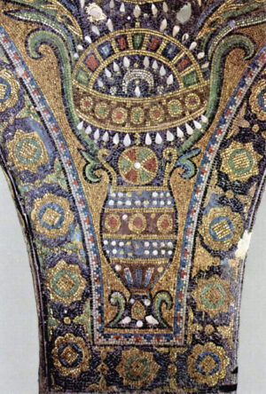 Photograph of an ancient mosaic detail featuring a central ornate vase with geometric patterns in gold, red, blue, and white tiles. Surrounding the vase are intricate floral and circular motifs in green, gold, and black, highlighting detailed craftsmanship and decorative style.
