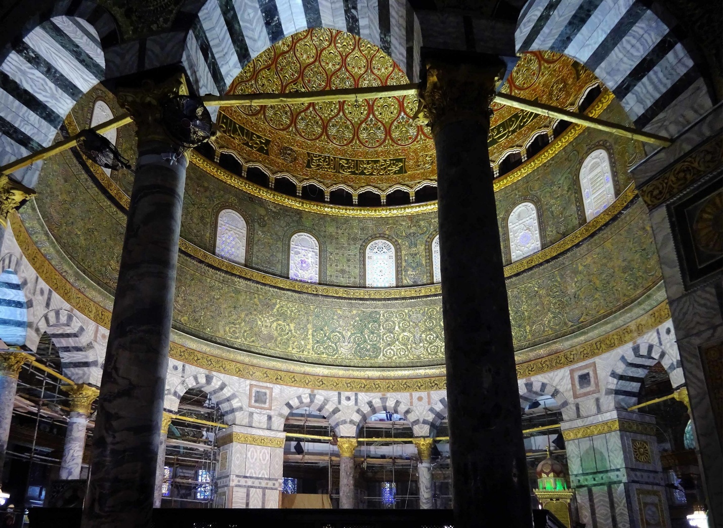 Photograph of Interior view of the Dome of the Rock dome featuring intricate gold and green mosaic patterns with arched windows and striped black-and-white columns. The image highlights architectural details of a historic religious structure, showcasing craftsmanship and decorative elements in a circular layout.