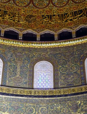 Photograph of an ornately decorated interior wall and dome section featuring intricate Islamic calligraphy and geometric patterns in gold, blue, and green hues. The design includes arched windows and detailed floral motifs, highlighting traditional Islamic architectural artistry.