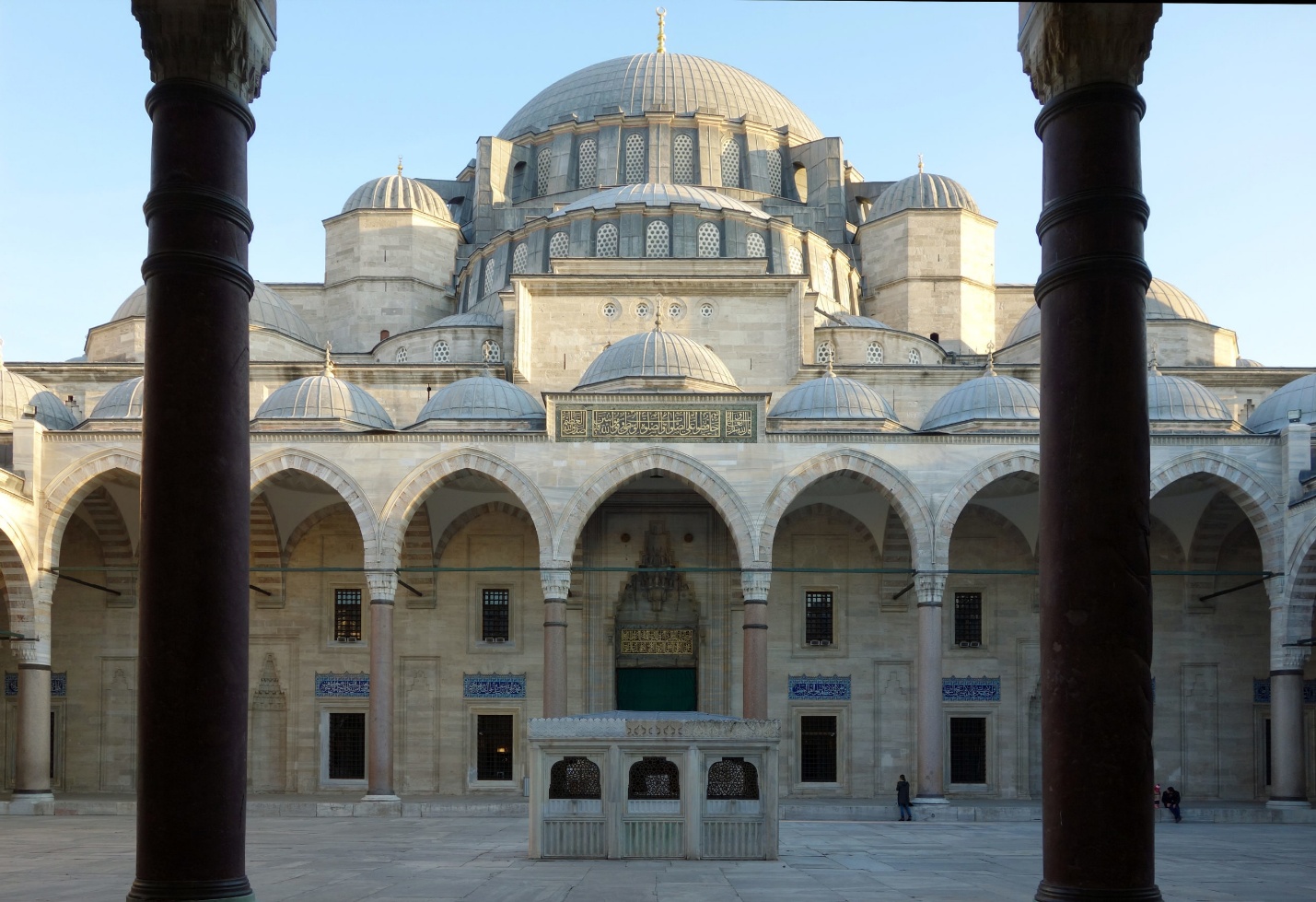 Photograph of a historic mosque courtyard featuring multiple domes and arched colonnades framed by two dark columns in foreground. Central ablution fountain and intricate stonework highlight Ottoman architectural style under clear blue sky.