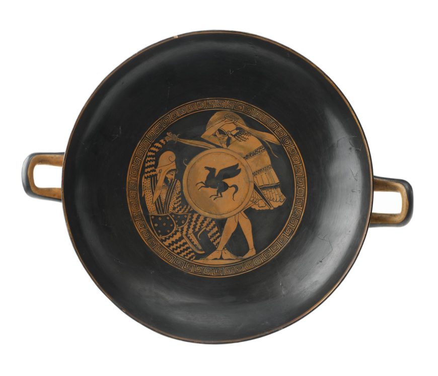 Photograph of an ancient Greek black-figure kylix, a type of drinking cup, featuring a central circular design with two warriors in combat. The artwork includes detailed armor, helmets, and a shield with a pegasus emblem, framed by a decorative geometric border.