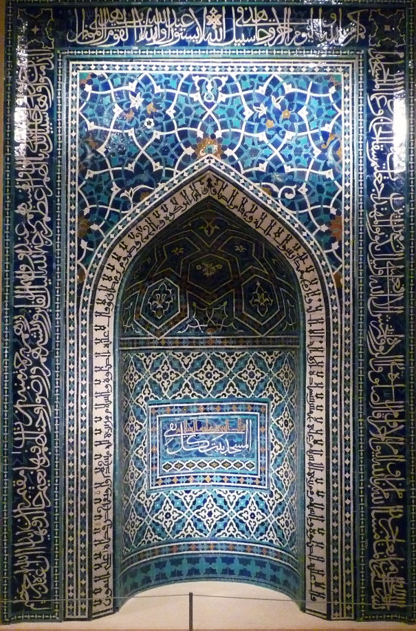 Photograph of an ornate Islamic architectural niche decorated with intricate blue and turquoise tilework featuring geometric patterns and Arabic calligraphy. The design includes a pointed arch framed by multiple bands of calligraphy in white script on dark blue backgrounds, highlighting religious or poetic inscriptions.