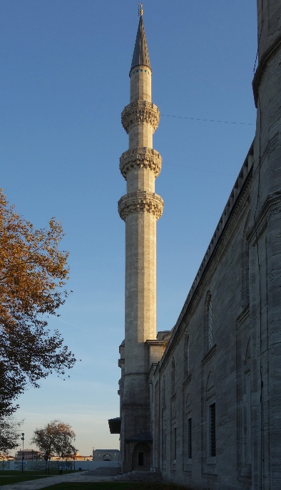 Photograph of a tall, slender minaret attached to a stone mosque building under clear blue sky. The minaret features three ornate balconies and a pointed spire, with autumn trees visible on the left side.