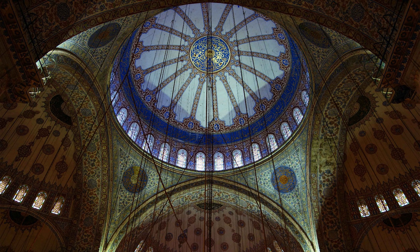 Photograph of an ornate mosque dome showcasing intricate blue and gold Islamic geometric patterns and calligraphy. The dome features a central circular design surrounded by radiating lines, with arched windows allowing natural light to illuminate detailed wall decorations below.