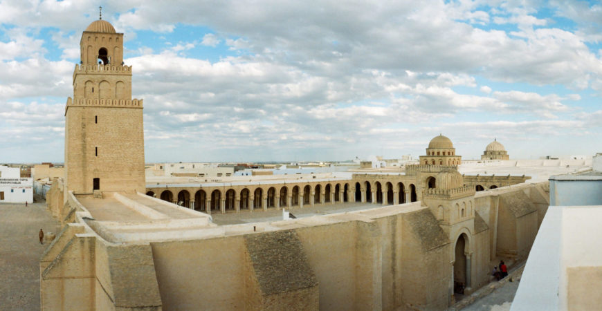 Photograph of a large historic mosque courtyard featuring a tall minaret on the left and arched colonnades surrounding the open space. The structure is made of beige stone with a clear blue sky and scattered clouds above, highlighting architectural details and expansive layout.