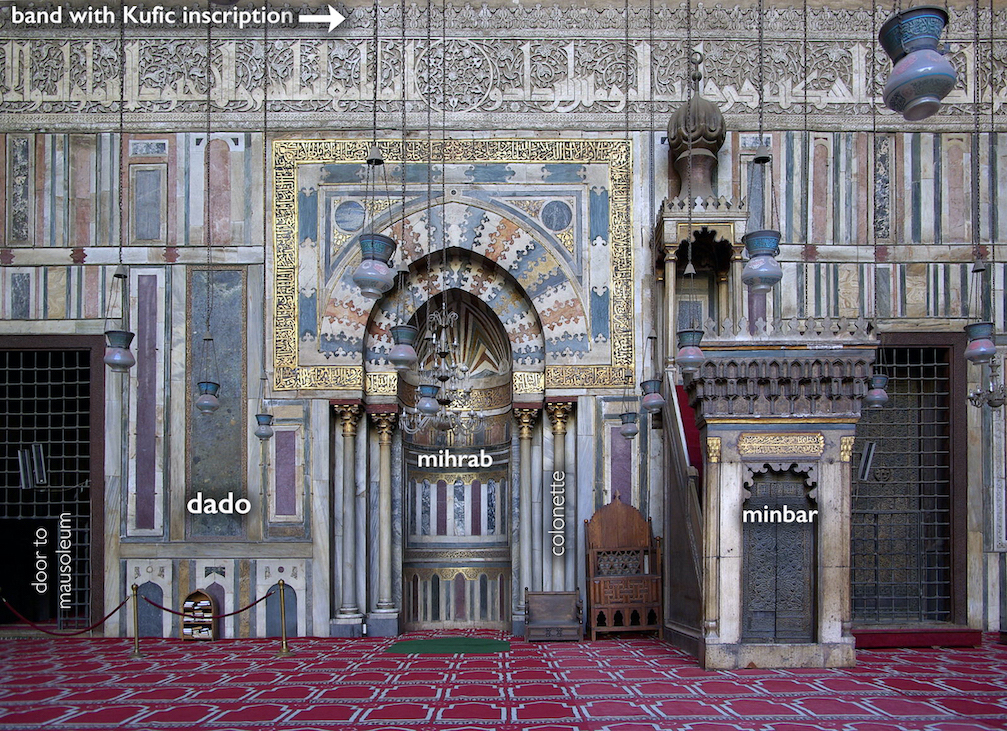 Photograph of an ornately decorated mosque interior featuring labeled architectural elements including a mihrab, minbar, dado, colonette, and door to mausoleum. The walls display intricate geometric patterns, a band with Kufic inscription, and hanging lamps, highlighting Islamic art and religious design.