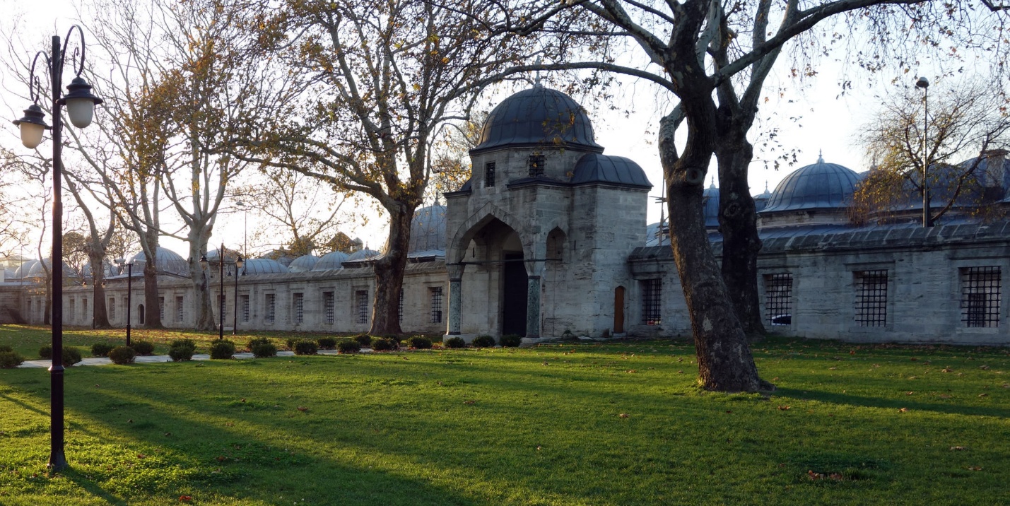 Photograph of a historic stone building with domed roofs and an arched entrance, surrounded by leafless trees and a green lawn. The scene is illuminated by soft sunlight, casting long shadows and highlighting architectural details and a vintage streetlamp on the left.
