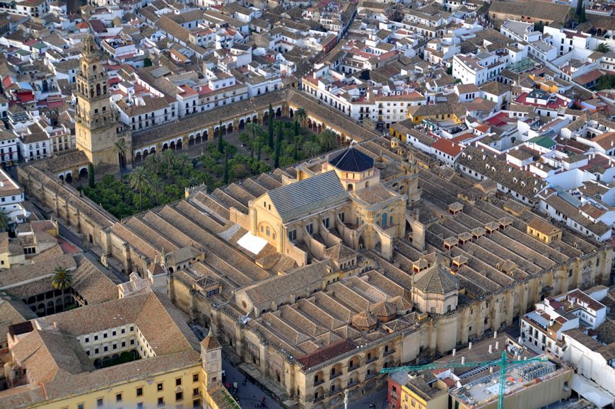 Aerial photograph of a large historic mosque-cathedral complex surrounded by dense urban buildings with white walls and tiled roofs. The structure features a prominent bell tower, multiple roof sections with varying heights, and an inner courtyard with greenery.