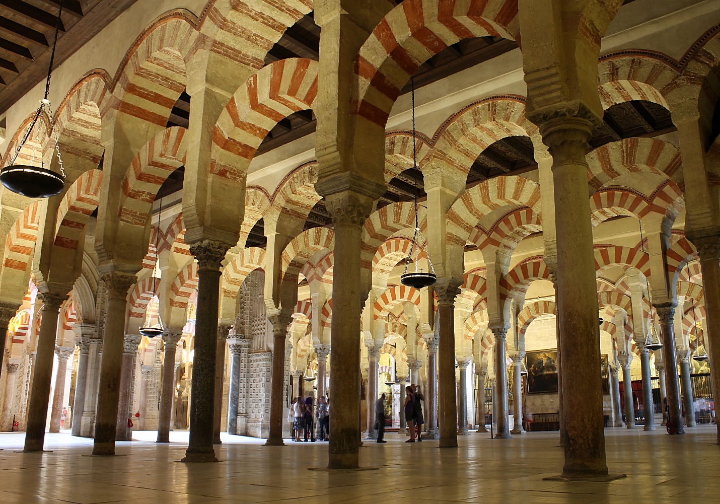 Photograph of a historic mosque interior featuring rows of horseshoe arches with alternating red and white voussoirs supported by stone columns. The architectural design highlights Islamic art and engineering, with natural light illuminating the expansive prayer hall and visitors exploring the space.