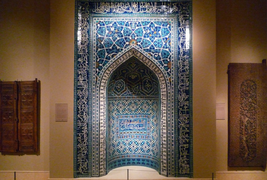 Photograph of a large, ornate Islamic Mihrab Niche tile panel featuring intricate blue and turquoise geometric patterns and Arabic calligraphy framing a pointed arch niche. The panel is displayed on a beige wall between two other wooden and carved artifacts, highlighting traditional Islamic art and architectural decoration.