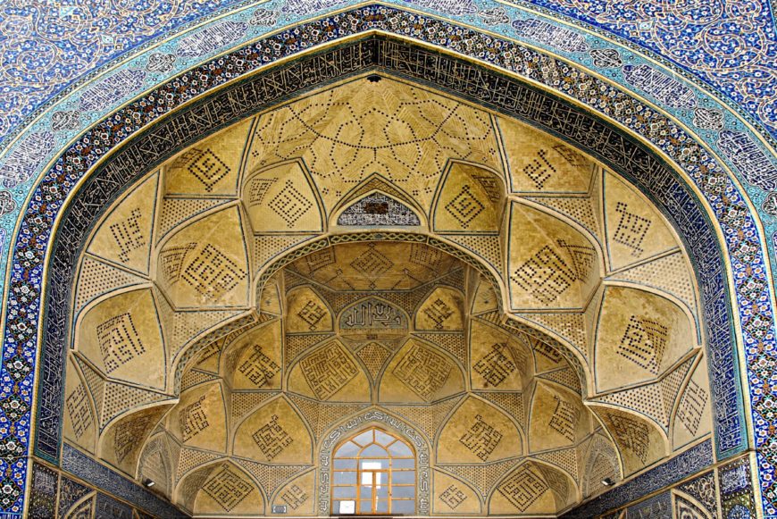 Photograph of an ornate Islamic architectural archway featuring intricate geometric patterns and calligraphy in blue and gold tones. The arch includes a central window and detailed tile work with symmetrical designs and Arabic script.
