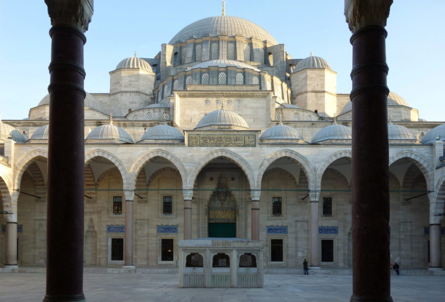 Photograph of a historic mosque courtyard featuring multiple domes and arched colonnades framed by two dark columns in foreground. Central ablution fountain and intricate stonework highlight Ottoman architectural style under clear blue sky.