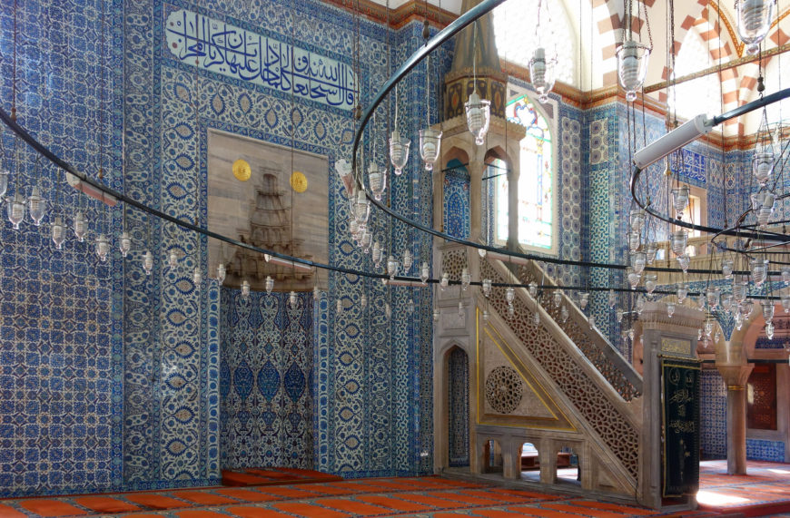 Photograph of an ornately decorated mosque interior featuring intricate blue and white tilework with Arabic calligraphy and a large wall painting of a sailing ship. The space includes a carved stone minbar (pulpit), stained glass windows, and hanging circular chandeliers with glass lamps, highlighting traditional Islamic architectural elements.