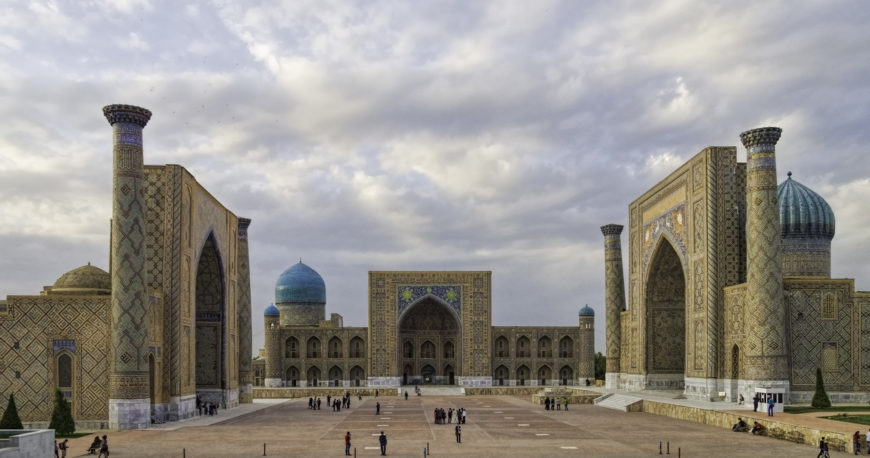 Photograph of Registan Square in Samarkand, Uzbekistan, showcasing three grand madrassas with intricate tilework and large arched entrances under a cloudy sky. The image highlights the central courtyard with scattered visitors, emphasizing architectural symmetry and vibrant blue domes.