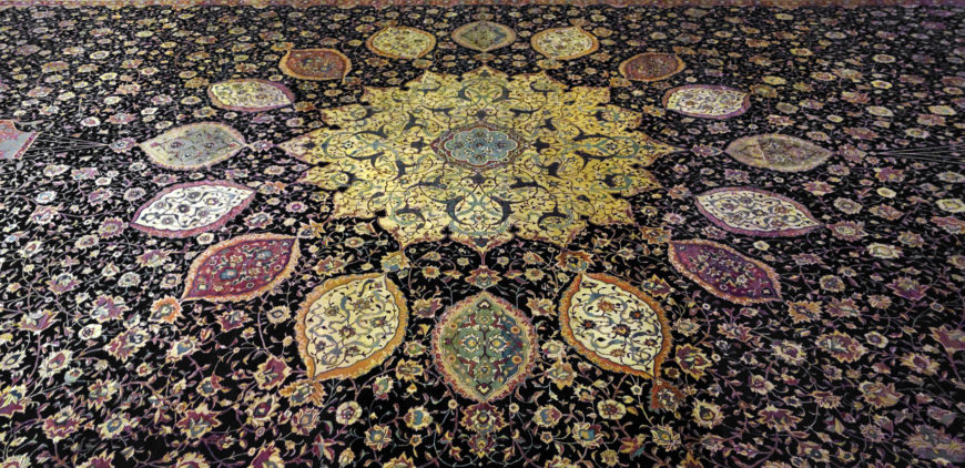 Photograph of an intricately patterned carpet featuring a central circular floral motif in gold and green hues, surrounded by petal-shaped designs in various colors including red, purple, and beige. The carpet's background is dark with dense floral patterns in pink and beige.