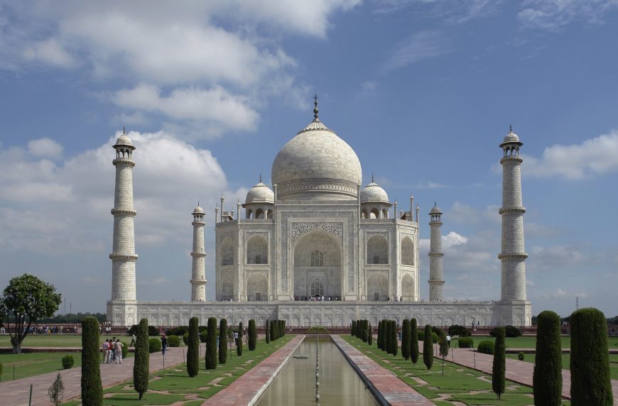 Photograph of Taj Mahal, a white marble mausoleum in India, showcasing its central dome flanked by four minarets under a partly cloudy sky. Foreground features a reflecting pool and symmetrical garden pathways leading to the monument, highlighting its architectural symmetry and intricate design.