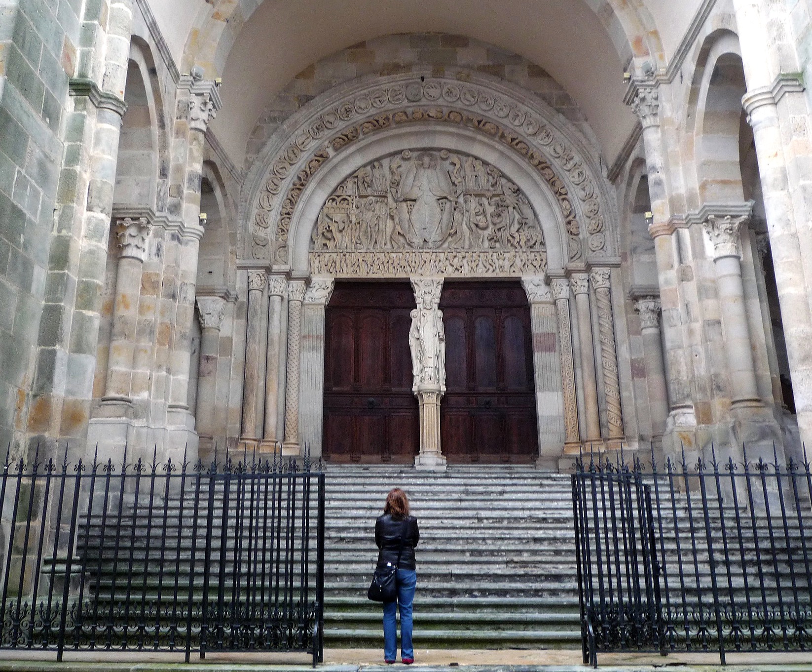 Photograph of a large, ornate church entrance featuring a wide staircase, dark wooden double doors, and an intricately carved stone archway with religious figures and decorative patterns. A person stands at the base of the steps facing the entrance, highlighting the grand scale and detailed architectural elements of the historic structure.