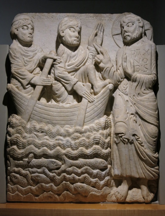 Bas-relief sculpture depicting three figures, two in a boat with oars and one standing on water, holding a book inscribed with Greek letters. Carving features detailed waves and fish below boat, with draped clothing and halos indicating biblical context.