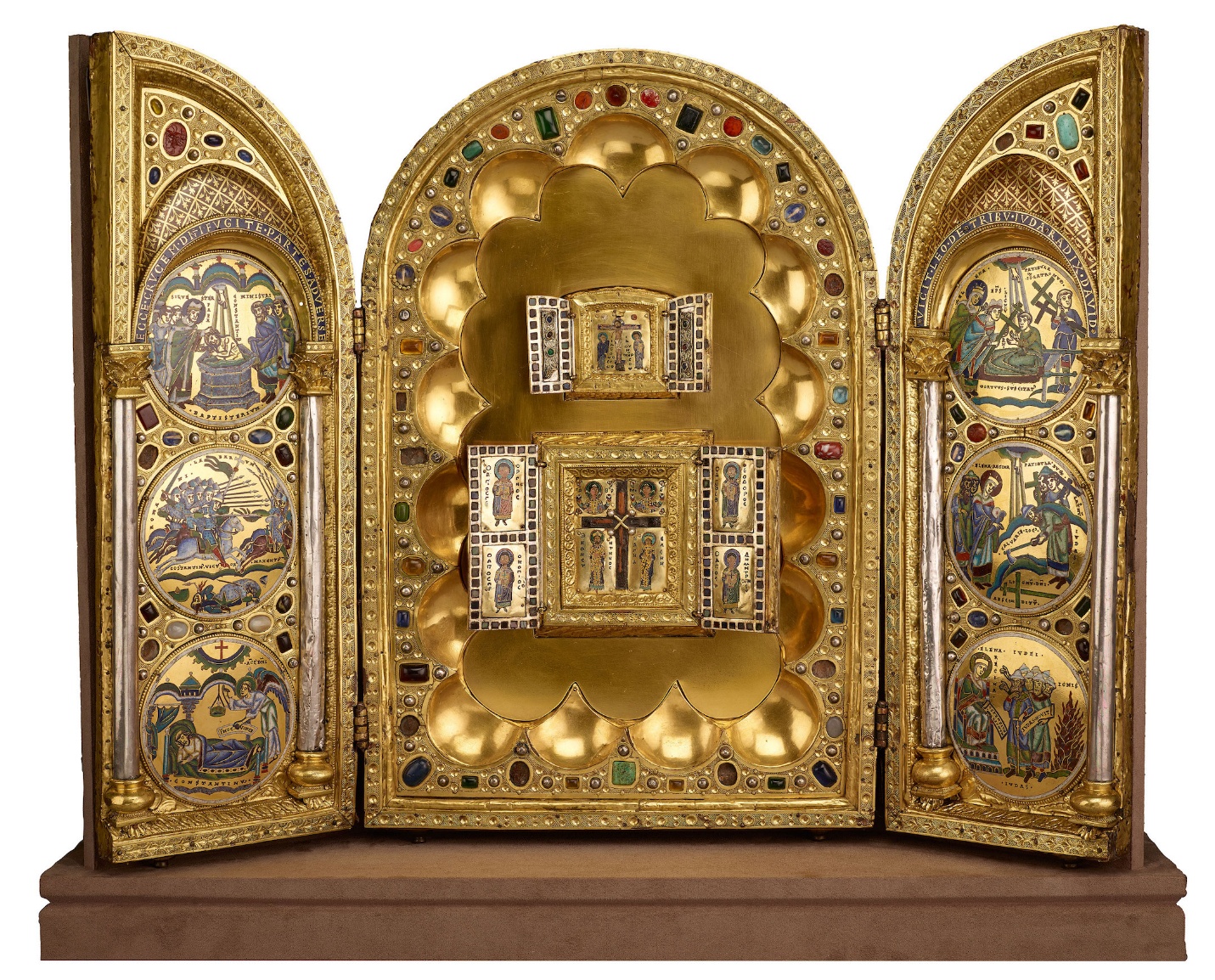 A photograph of an ornate triptych altarpiece featuring gold panels adorned with colorful gemstones and intricate religious iconography. Central panel displays a crucifixion scene with hinged doors revealing additional miniature religious figures, while side panels contain circular painted biblical scenes framed by elaborate gold designs.