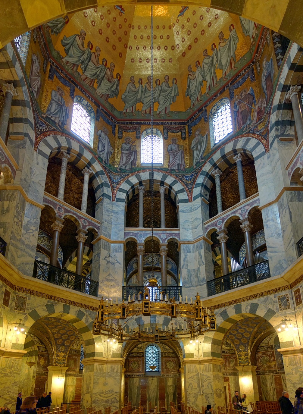 Photograph of an ornate cathedral interior showcasing a high vaulted ceiling adorned with religious frescoes and intricate mosaics in gold and blue tones. Architectural features include arched windows, marble columns with striped patterns, and a large circular chandelier hanging above visitors admiring the historic space.