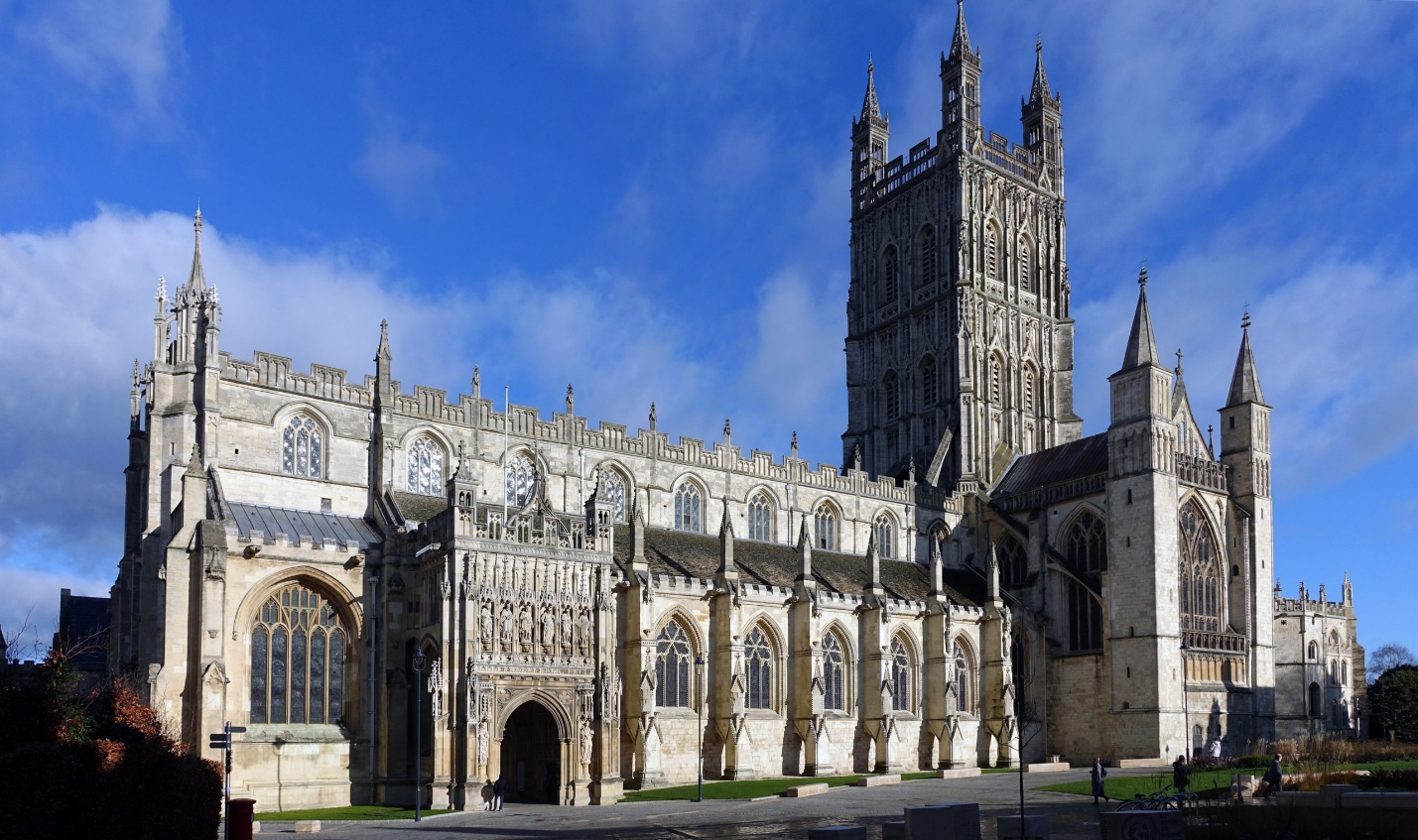 Photograph of a large Gothic-style cathedral with intricate stonework, tall spires, and a prominent central tower under a bright blue sky. The structure features arched windows, detailed carvings, and a well-maintained lawn in the foreground, highlighting historic architectural design.