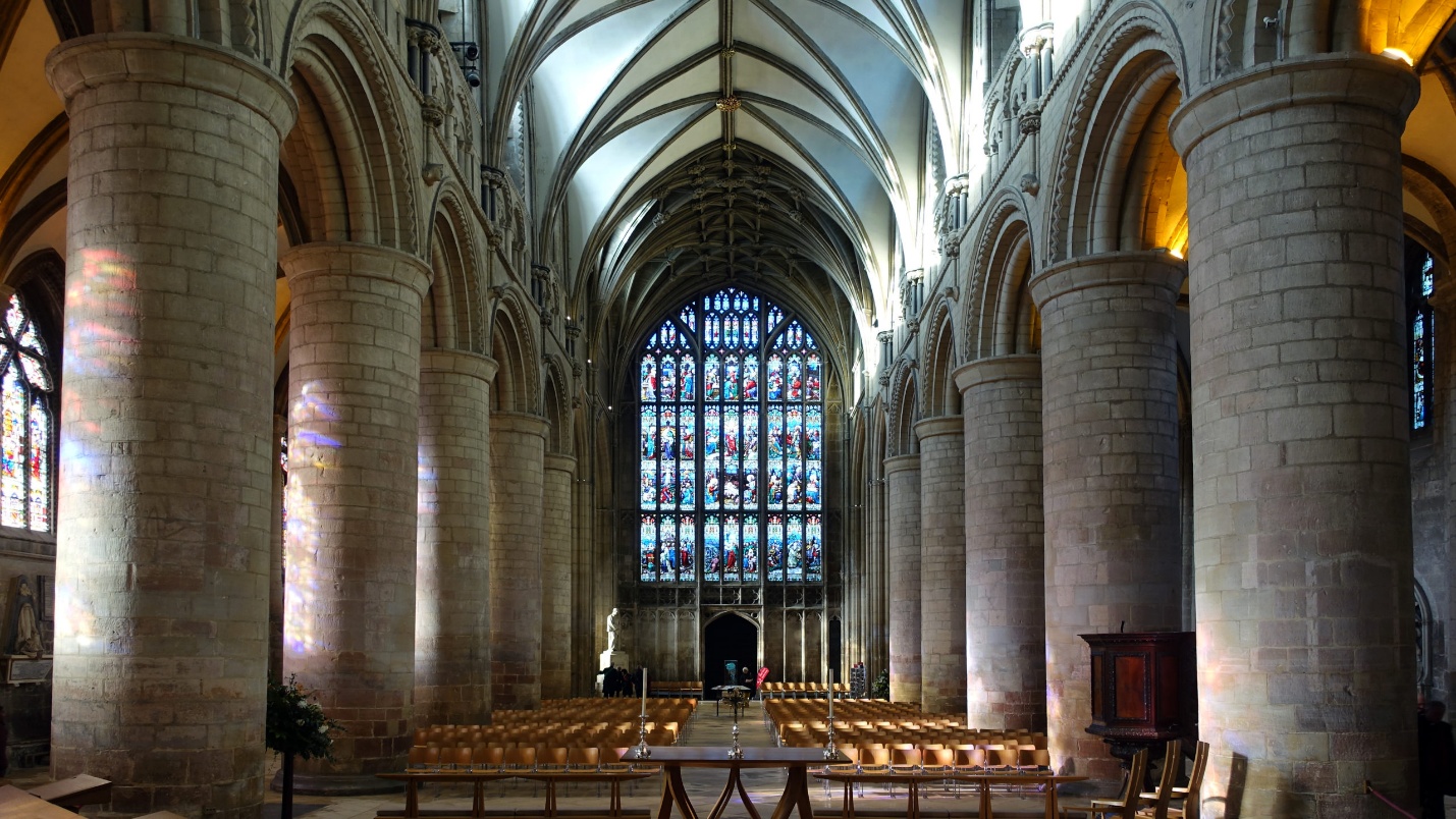 Photograph of a cathedral interior showcasing tall stone columns and vaulted ceilings with intricate arches. Stained glass windows at the far end allow colorful light to illuminate rows of wooden pews, highlighting architectural grandeur and historical design.