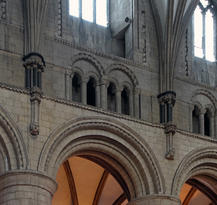 Photograph of Romanesque architectural details inside a historic stone building, featuring large rounded arches supported by thick columns and small paired windows above. The structure highlights intricate stone carvings, including decorative capitals and sculpted faces on column bases, with natural light filtering through tall windows.