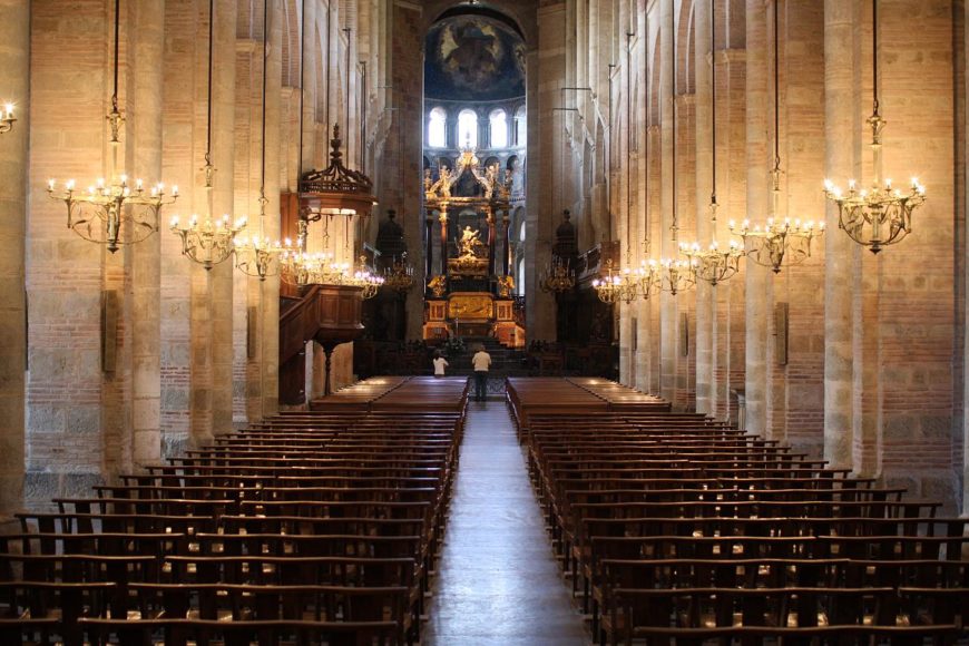 Photograph of a large, historic church interior featuring rows of wooden pews aligned along a central aisle leading to an ornate altar with golden decorations. Warm lighting from chandeliers and wall sconces highlights tall stone columns and high vaulted ceilings.